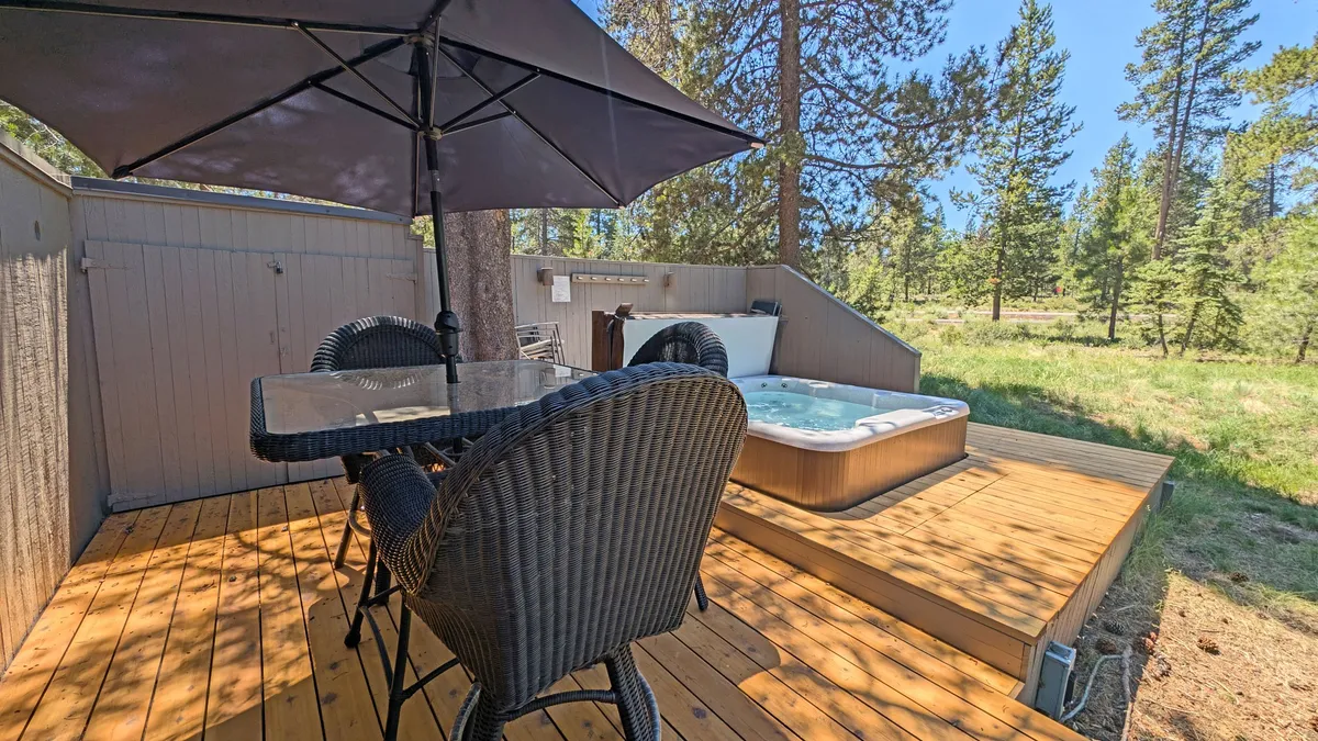 This image showcases a spacious outdoor deck featuring a large dining table and a hot tub, surrounded by tall trees. The setting is serene and inviting, ideal for outdoor meals, relaxation, or soaking in the hot tub while enjoying the surrounding natural beauty.