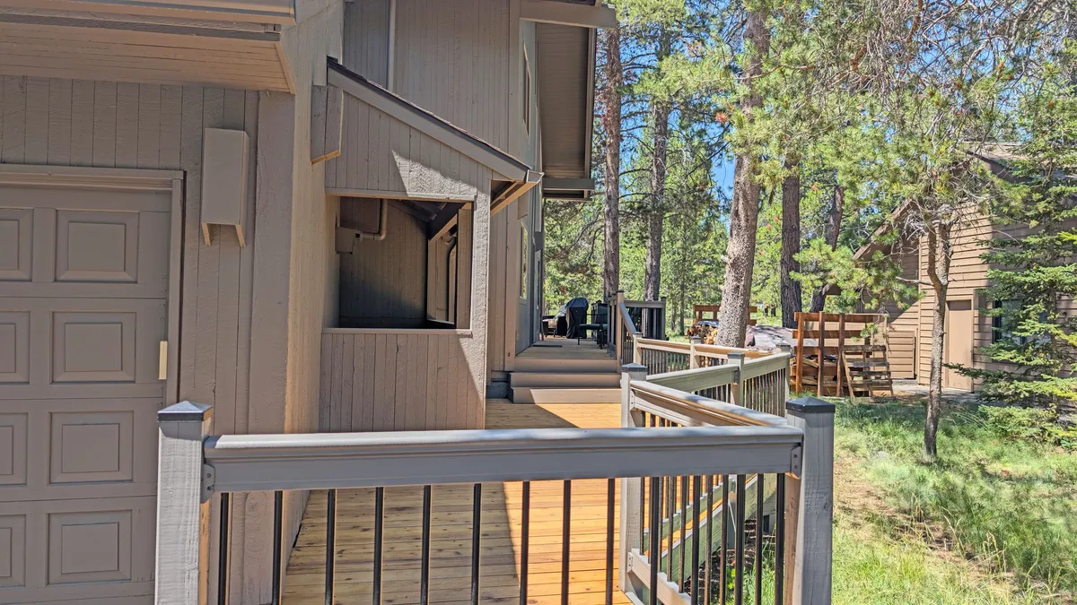 This image shows the exterior of Indian 3, featuring a wooden deck with sturdy railings, the garage door (where one of the EV chargers is located), and a backdrop of tall, surrounding trees. The deck suggests a seamless transition from home to nature, with space for relaxation and outdoor enjoyment.