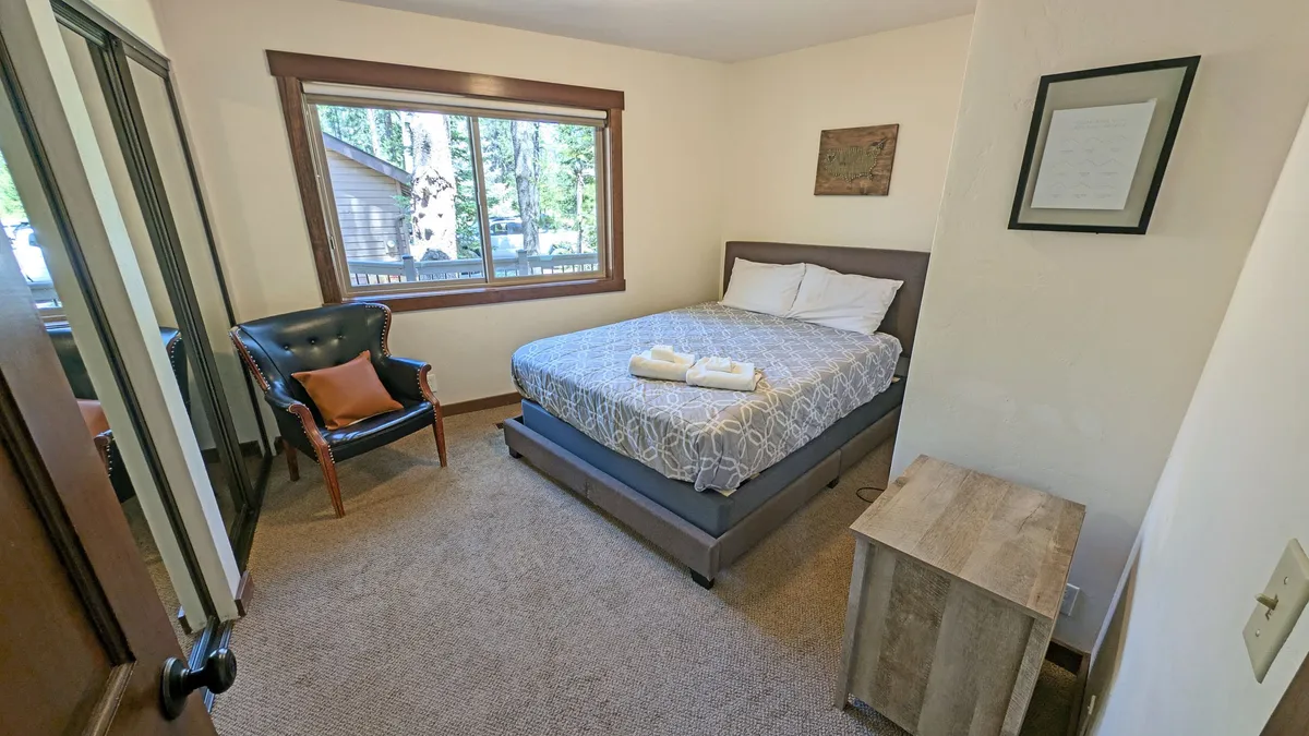 A queen bed with patterned bedding is positioned near a window that frames views of surrounding trees. An armchair with an accent pillow sits beside a nightstand, creating a cozy reading nook. The room is bathed in soft, natural light, emphasizing a calm and serene atmosphere.