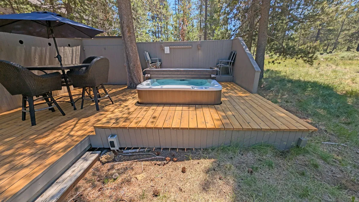 This image features a spacious outdoor deck with a bubbling hot tub, a dining table, and comfortable outdoor seating surrounded by lush trees. The setting is tranquil and inviting, with ample room for gatherings, relaxation, and enjoying meals in the fresh air. The deck showcases both the hot tub and al fresco dining options, encapsulating the essence of outdoor resort living.