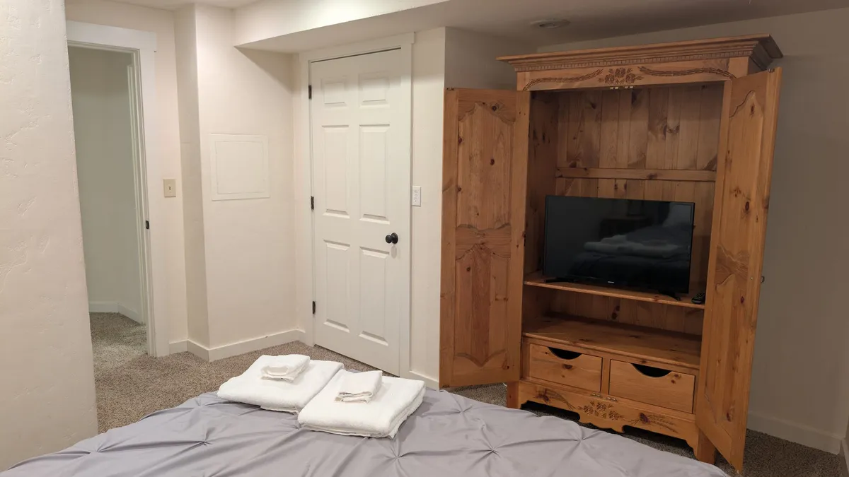 A queen bedroom with a comfortable bed draped in gray bedding, a wooden armoire that houses a TV, and neatly folded towels on the bed—offering a cozy spot for relaxation and movie nights.