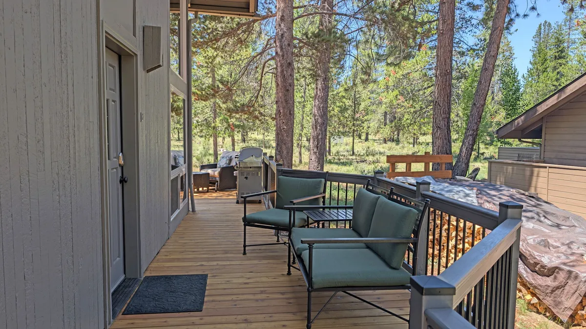 A serene section of the wooden deck is shown with green chairs arranged for relaxation, surrounded by tall trees and featuring a grill area perfect for hosting BBQs. The space invites guests to unwind, entertain, and savor the outdoors, with sunset views and stargazing opportunities suggested by the open sky and forest backdrop.