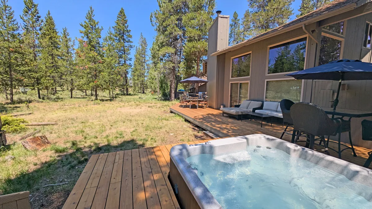 A spacious wooden deck set amidst tall pine trees, featuring a private hot tub, comfortable outdoor seating, and plenty of open space for relaxing after a day of adventure. The deck feels inviting and surrounded by nature, making it the perfect spot for unwinding under the stars or sharing stories with friends after exploring local trails.