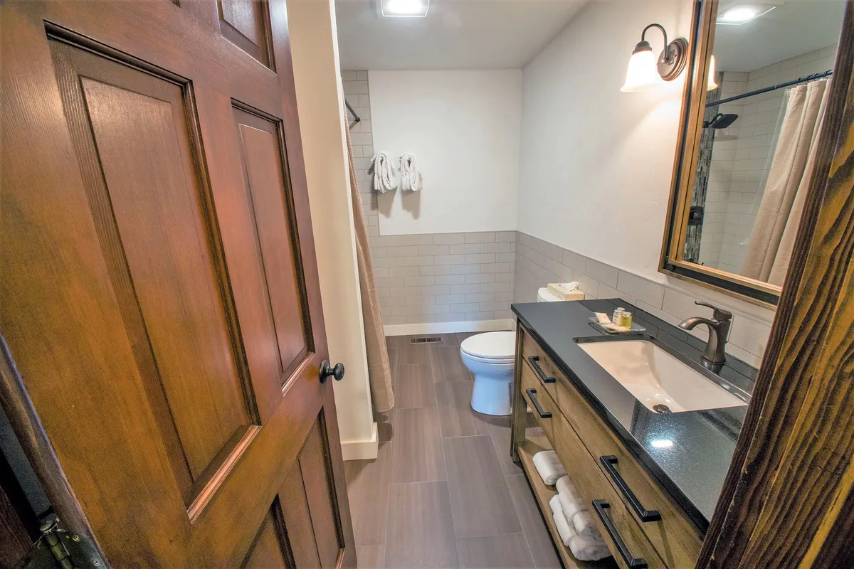 A refreshing bathroom space featuring a wooden door, vanity with a sleek black countertop, a large mirror, toilet, and crisp towels hanging on the wall. The room feels spa-like and is ideal for morning routines and relaxation.