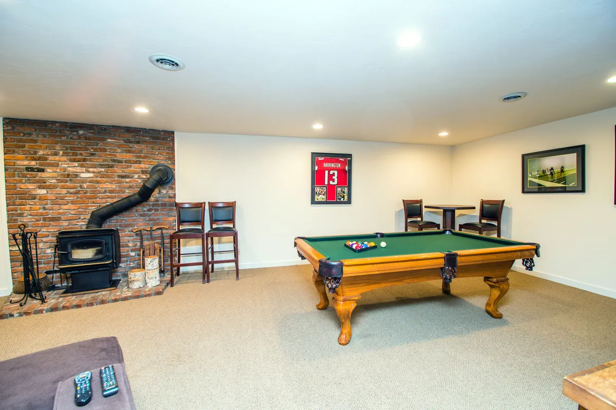 A spacious entertainment room featuring a pool table as the centerpiece, with a rustic brick wall behind it, bar stools for extra seating, and a wood stove that adds to the cozy atmosphere. The area is designed for socializing, playing games, and relaxing with friends or family.