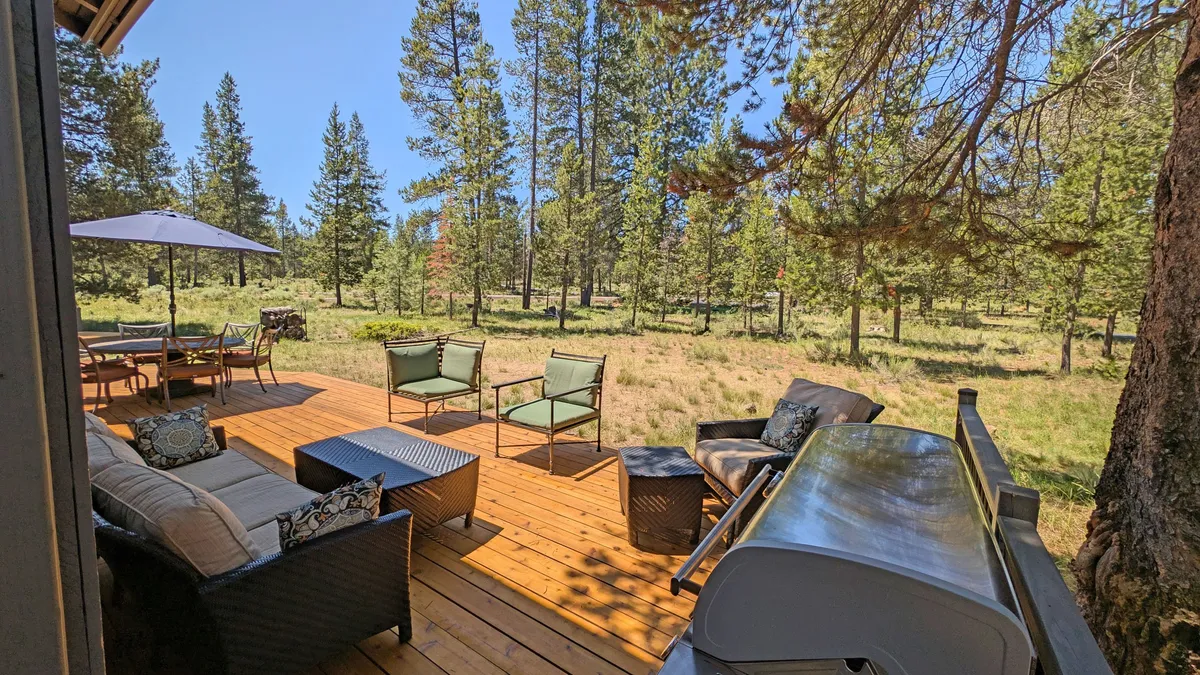 A tranquil exterior view of the spacious wrap-around deck surrounded by lush trees. The deck features comfortable seating and a grill, offering plenty of room to relax, socialize, or enjoy a meal outdoors. The open space and connection to nature are emphasized, suggesting ample room for pets and family to enjoy the setting together.