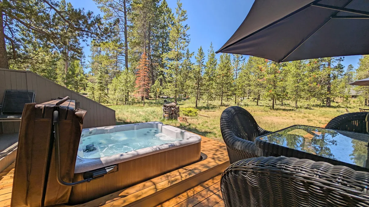 The image captures a hot tub nestled on the deck, surrounded by comfortable outdoor seating and towering trees. A fire pit adds extra warmth and ambiance for cozy evenings. The setting is peaceful, ideal for soaking under the stars, socializing, or enjoying a quiet morning coffee as sunlight filters through the trees.