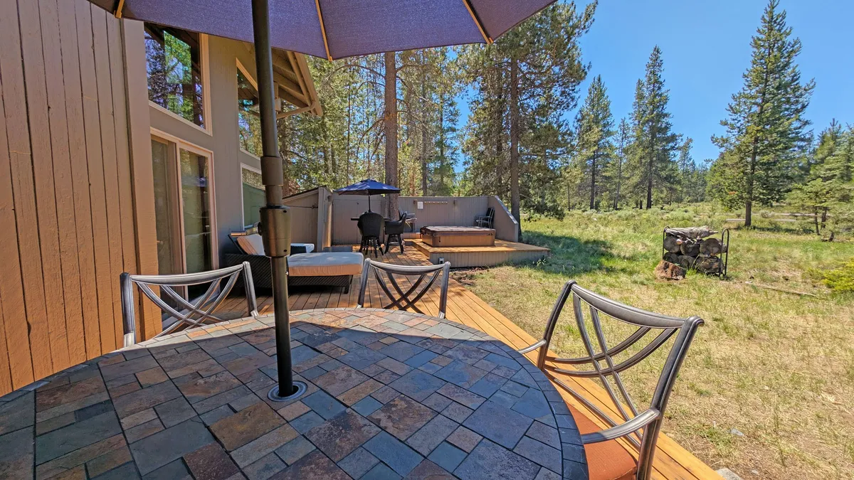 A welcoming outdoor patio with a table and umbrella, comfortable seating area, and a fire pit, all set amidst lush trees. The arrangement encourages group dining, evening relaxation, and cozy gatherings around the fire pit.