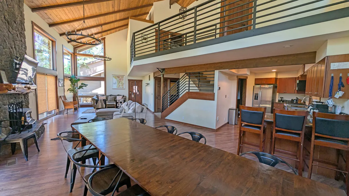 The image showcases a spacious, sunlit interior with a long wooden dining table at the center, perfect for large gatherings. Large windows flood the room with natural light, revealing views of the outdoors, while a beautiful stone fireplace adds warmth and charm. A staircase leads to the upper level, offering a glimpse of the home's generous space and inviting layout.