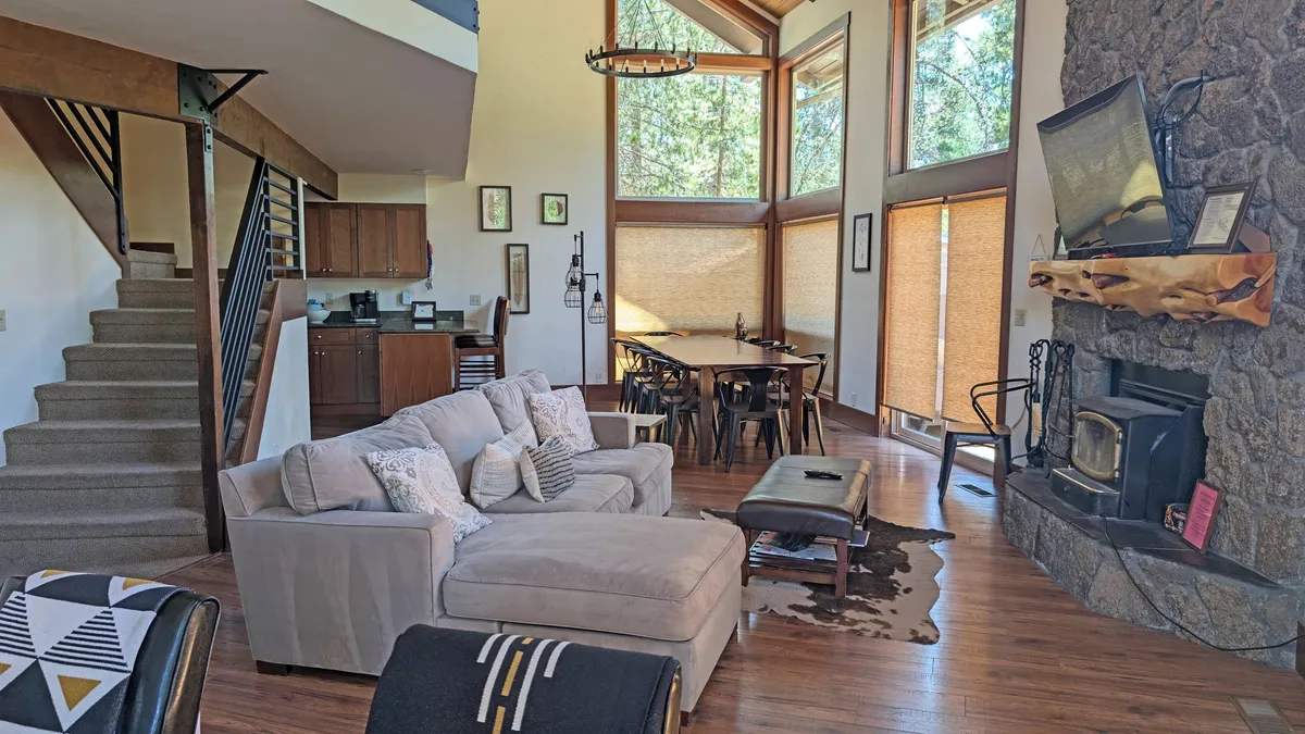 A sunlit living room with large windows that let in natural light, a stone fireplace, a comfortable sectional sofa, and a dining area. The atmosphere is relaxed and open, encouraging conversation, relaxation, and informal meetings.